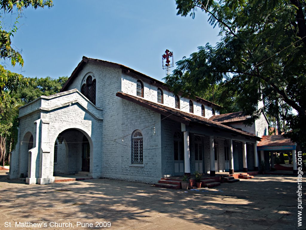Exterior view of St. Matthew's Church in Pune, a light grey stone building with arched windows and a red neon cross on the roof, surrounded by trees.